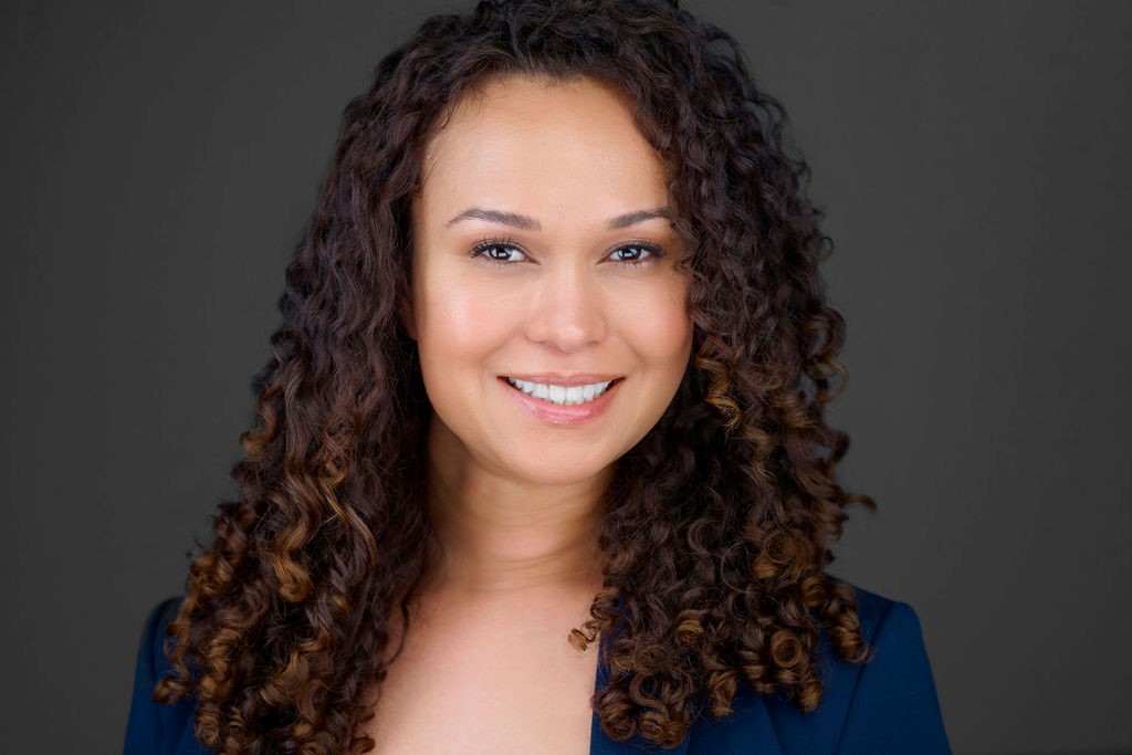 Smiling person with curly hair wearing a dark blazer against a gray background.